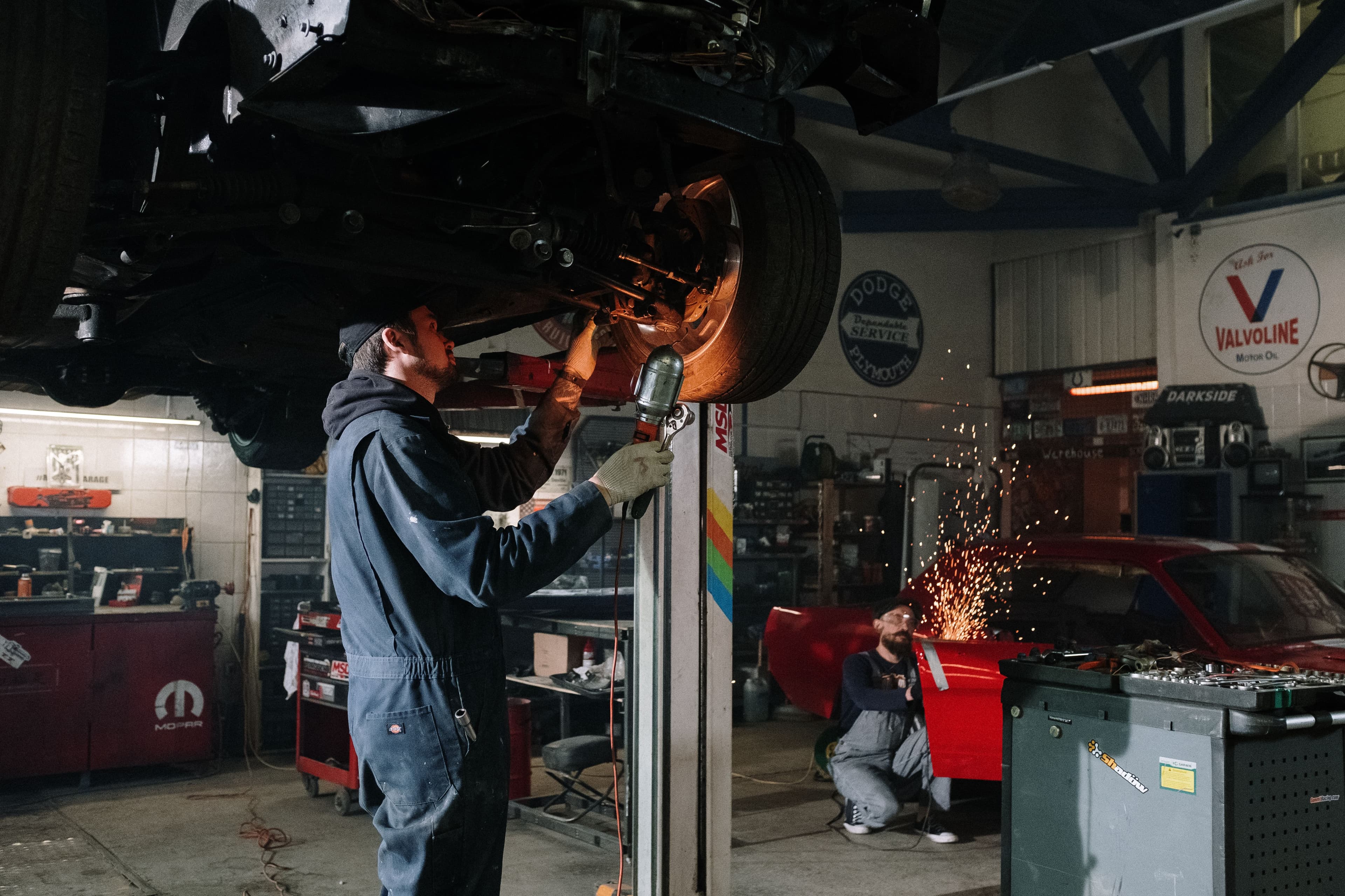 Mechanici při práci na voze — jeden u podvozku na zvedáku, druhý u broušení s jiskrami